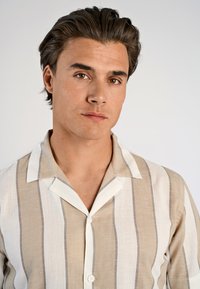 Jongetje met donker haar, gekleed in een beige en witte horizontaal gestreepte overhemd met kraag, kijkt recht vooruit met een neutrale uitdrukking.