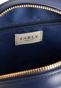Pochette zippée bleu marine avec quincaillerie à fermeture éclair dorée ; la doublure intérieure présente une texture lisse. L'étiquette indique "FABLE ENGLAND" en texte noir.