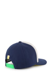 Navy pet met een witte zachte bontpaneel, voorzien van een groene verstelbare band aan de achterkant en een gele accentlabel. Plat vizier ontwerp.