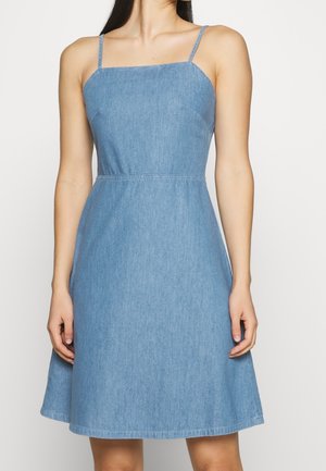 Kvinde iført ærmeløs, lyseblå denimkjole med tynde stropper og A-formet nederdel, stående mod en ensfarvet lys baggrund.