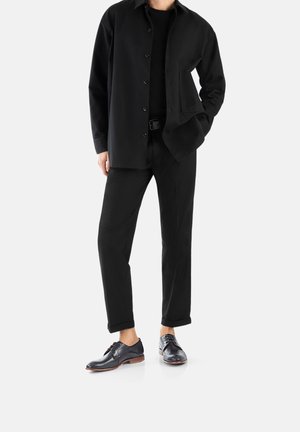 Chemise noire, T-shirt noir et pantalon noir, associés à des chaussures noires brillantes avec un motif texturé et une semelle marron.