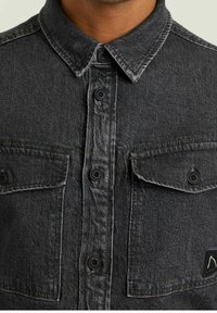 Camicia in denim nero con chiusura frontale a bottoni, due tasche sul petto, colletto a punta e bottoni neri a contrasto; texture leggermente scolorita.