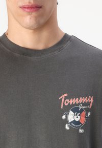 Šedé bavlněné tričko s kulatým logem a kreslenou postavičkou v červené, bílé a modré barvě, doplněné textem "Tommy Jeans Records."