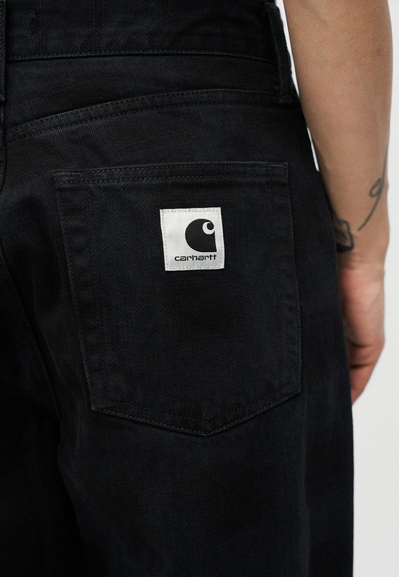 Jeans neri in denim con finitura testurizzata, dotati di una tasca posteriore e di un'etichetta quadrata bianca con un logo nero.