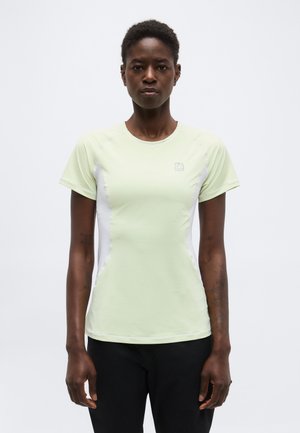 Femme aux cheveux courts portant un t-shirt de sport à manches courtes vert clair et un pantalon noir, debout devant un fond blanc uni.