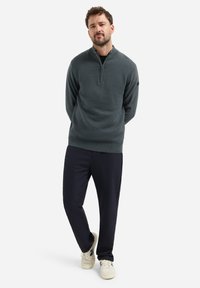 Man met een baard die een donkergroene geribbelde half-zip trui, zwarte broek en witte sneakers draagt, staande met de handen achter zijn rug.