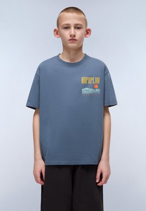 Camiseta de algodón gris con mangas cortas, presenta un estampado gráfico que incluye el texto "NAPAPIJRI" y un sol, combinada con pantalones cortos sueltos de color negro.