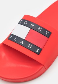 Piros csúszópapucs széles pánttal, amelyen egy fekete téglalap alakú folt található, fehér betűkkel a "TOMMY JEANS" márkanevével.
