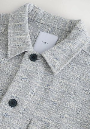 Camisa de patrón azul claro y blanco con cuello, tela texturizada y dos botones oscuros cerca del cuello. La etiqueta dice "NEXT."