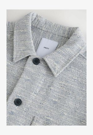 Hellblaues und weißes gemustertes Hemd mit einem kragenförmigen Ausschnitt, strukturiertem Stoff und zwei dunklen Knöpfen in der Nähe des Kragens. Das Etikett trägt die Aufschrift "NEXT."