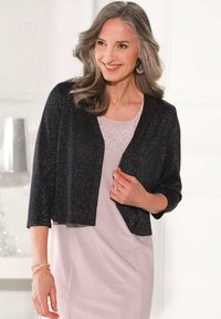 Schwarze, glitzernde Strickjacke mit Dreiviertelärmeln, getragen über einem hellrosa Kleid mit dezenten Verzierungen. Einfache runde Ausschnitt.