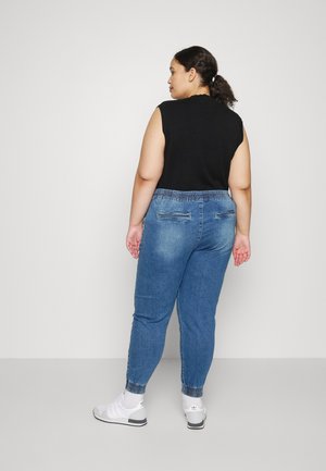 Vrouw met donker haar in een paardenstaart, die een zwart mouwloos topje draagt, blauwe jogger jeans, witte sokken en witte sneakers, staat met haar rug naar de camera.