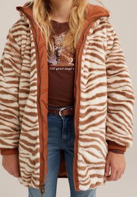 Hnědý a krémový pruhovaný umělý kožich se zipem v přední části, žebrovanými manžetami a oranžovou podšívkou, nošený přes hnědou košili a džínové kalhoty.