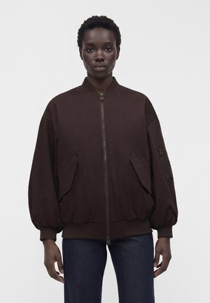 Osoba s krátkymi vlasmi oblečená v tmavohnedú oversized bomber bundu s prednými vreckami a tmavými nohavicami, stojaca pred jednofarebným pozadím.