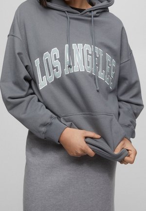 Personne portant un sweat à capuche gris avec le texte « LOS ANGELES », tirant sur le tissu à l'avant, assorti à une jupe ou un pantalon gris.