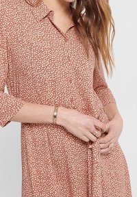 Ruskea kuvioitu mekko napitus edessä, lyhyillä hihoilla ja vyötäröllä solmittava yksityiskohta. Kangas vaikuttaa kevyeltä ja teksturoidulta.