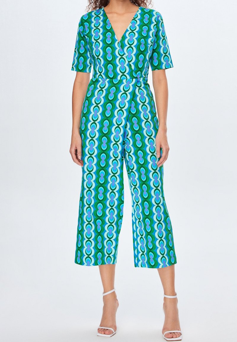 Femme portant une combinaison à motifs géométriques verts, bleus et violets, à manches courtes et jambes larges, assortie à des talons blancs à bride cheville.