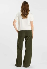 Una mujer con pelo largo y ondulado de color castaño está de espaldas, lleva una camisa blanca de manga corta y pantalones verdes olivo de pierna ancha.
