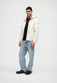 Κρεμ πανωφόρι φουσκωτό με κουκούλα, κλείσιμο με φερμουάρ και πλαινές τσέπες, φορεμένο πάνω από μαύρο πουκάμισο και συνδυασμένο με ανοιχτό μπλε τζιν και μαύρα παπούτσια.