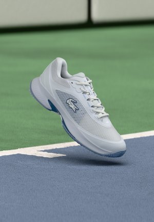Scarpa da tennis Lacoste bianca con suola e logo blu che fluttua sopra una superficie di campo da tennis blu e verde.