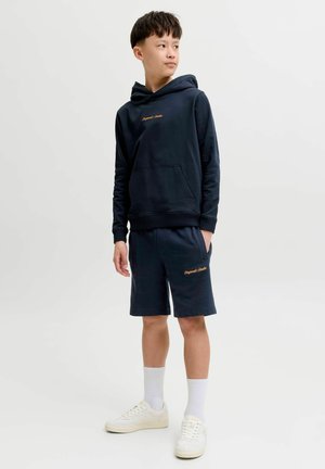 Jongen die een marineblauwe hoodie en korte broek draagt, met witte sokken en sneakers, staand en opzij kijkend tegen een effen achtergrond.