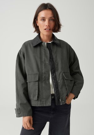 Femme aux cheveux courts et foncés portant une veste zippée vert foncé avec poches à rabat et un pantalon noir, debout devant un fond uni.