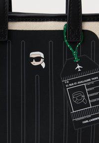 Saco tote com um exterior texturizado em preto, design riscado e um gráfico de uma pessoa com óculos de sol. Inclui uma etiqueta de bagagem verde.