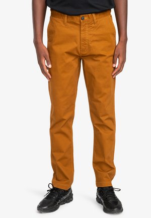 Pantaloni in cotone marroni con taglio dritto, chiusura frontale con bottone e dettagli di cucitura minimali. Abbinati a scarpe nere con lacci.