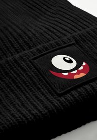 Black knit beanie featuring a textured design with a square patch displaying a playful monster face in white, red, and pink.