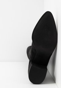 Botte en daim noir reposant semelle vers le haut contre un coin blanc, montrant la semelle texturée et un talon carré bas.