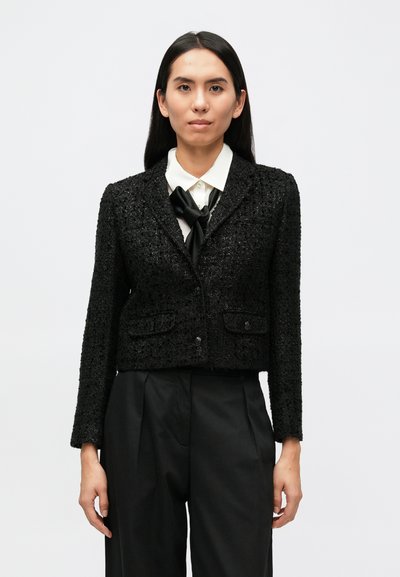 Zwart cropped blazer van een gestructureerde stof, met twee voorzakken en een enkele knoopsluiting. Gecombineerd met een wit overhemd en een strikje.