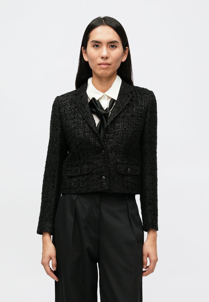 Fekete, vágott fazonú blézer texturált anyagból, két első zsebbel és egysoros gombolással. Fehér inggel és csokornyakkendővel párosítva.