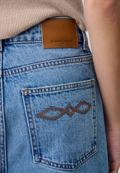 I jeans in denim azzurro chiaro presentano una tasca posteriore ben visibile con un motivo ricamato decorativo marrone e un'etichetta in pelle marrone.