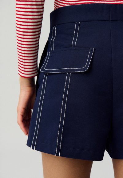 Shorts navy in cotone, con cuciture bianche e una tasca laterale. Vista ravvicinata che mostra una mano che afferra l'elastico.