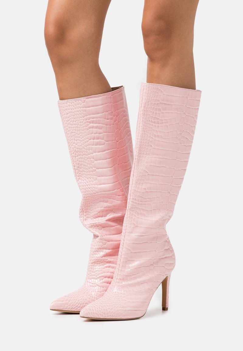 Jambes portant des bottes roses montantes à motif crocodile, bouts pointus et talons aiguilles hauts sur fond blanc.