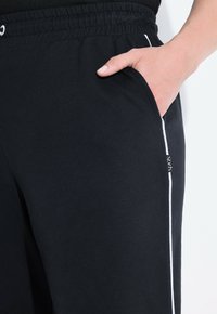 Schwarze Jogginghose mit weichem Material, ausgestattet mit einem weißen Seitenstreifen, einem elastischen Bund und einer Seitentasche. Markenlabel sichtbar.