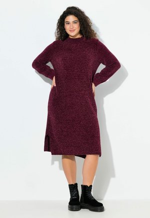 Robe en tricot bordeaux avec des manches longues, un col montant et des fentes latérales. Portée avec des bottes noires à lacets, mettant en valeur une coupe décontractée.