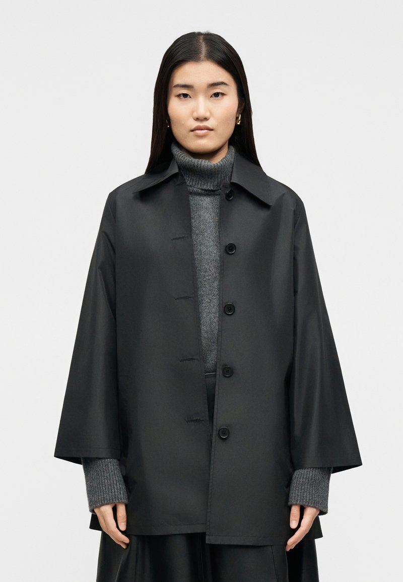 Γυναίκα που φοράει μαύρο παλτό με κουμπιά πάνω από γκρι πουλόβερ με ψηλό γιακά και μακριά μανίκια, σε απλό ανοιχτό φόντο.