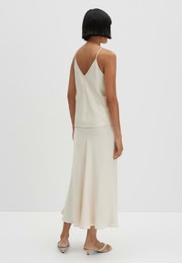 Cream-colored sleeveless dress with a V-back design, flowing skirt, smooth texture, and subtle seam details. Worn with light sandals.