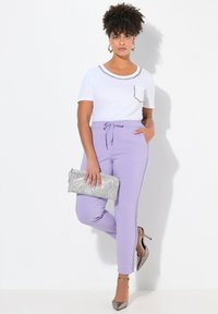 Vit T-shirt med ficka och dekorativ kedjedetalj, tillsammans med lila byxor och silverspikeklackar, hållande en metallisk kuvertväska.