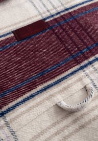 Pătură din lână cu un model tartan în bordeaux, crem și albastru. Dispune de un eticheta din piele și o mână pentru transport ușor.