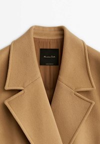 Col et revers d’un manteau en laine couleur camel avec une doublure marron visible et une étiquette noire « Massimo Dutti » à l’intérieur de la zone du cou.