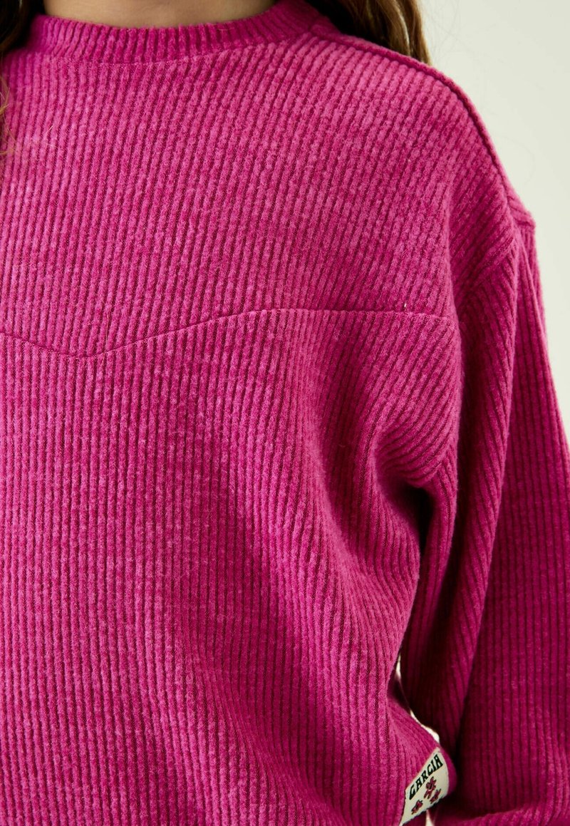 Gros plan sur une personne portant un pull côtelé rose vif à col rond avec une petite étiquette de marque blanche près de l'ourlet.
