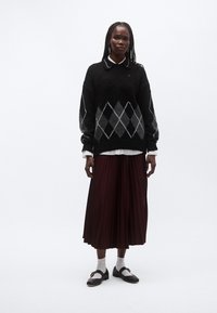 Maglione nero con motivo argyle bianco e grigio, indossato sopra una camicia bianca con colletto; gonna plissettata bordeaux; scarpe nere con calzini alla caviglia.