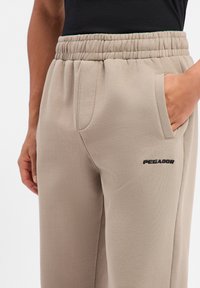Beige Jogginghose mit elastischem Bund, Seitentaschen und "PEGADOR" in Schwarz auf dem linken Oberschenkel gestickt. Weiche Stoffstruktur.
