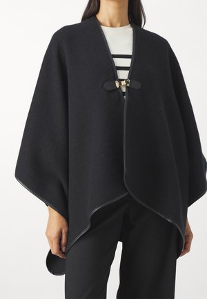 Femme portant une cape en laine noire avec bordure en cuir et fermoir doré, sur un haut blanc à rayures noires et un pantalon noir.