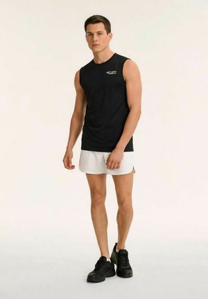 Junger Mann steht, trägt ein schwarzes ärmelloses Oberteil, weiße Shorts und schwarze Sportschuhe vor einem schlichten hellen Hintergrund.