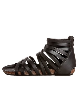 Black leather sandal with multiple crisscross straps, a flat sole, and a textured cork-like insert. Features a side zipper for adjustment.