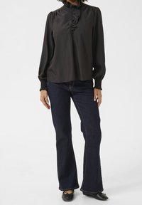 Blouse noire avec un devant froncé et un col haut, associée à un jean évasé bleu foncé. Le jean présente des coutures jaunes et une texture subtile.