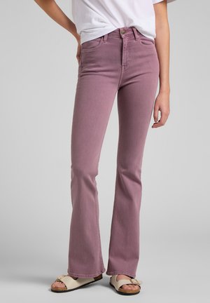 Personne portant un jean évasé taille haute violet clair, un t-shirt blanc et des sandales beige avec des semelles en liège sur un fond gris.